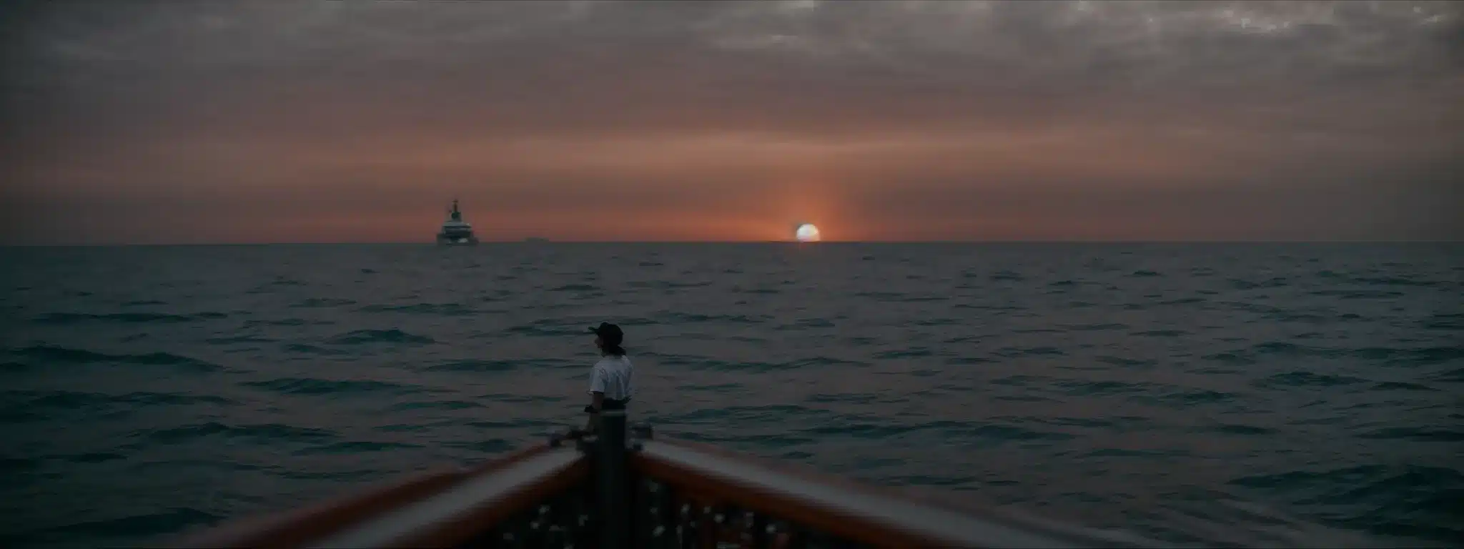 A Person Standing At The Helm Of A Ship, Navigating Through Calm Waters With A Sunset On The Horizon.