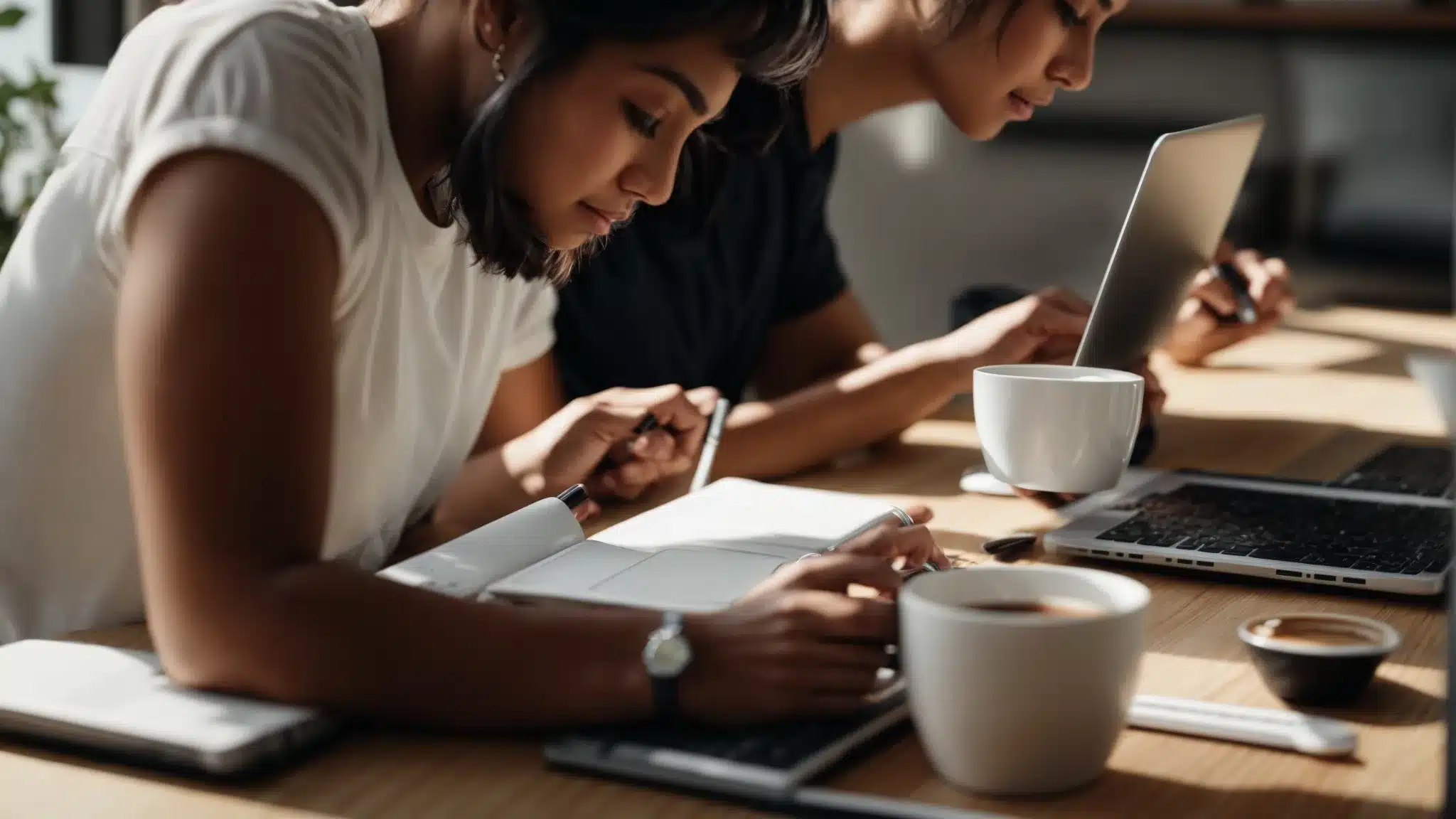 two people discuss over a laptop with a coffee cup and a notepad on the table, showing a search engine on the screen.